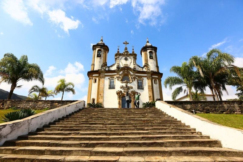 Igreja Nossa Senhora do Carmo em Ouro Preto vista de frente, no topo de uma longa escadaria com céu azul e palmeiras ao redor